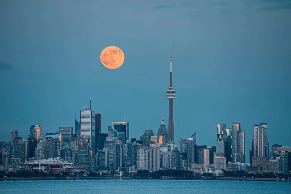 Toronto skyline at dusk with a full moon rising above the CN Tower, reflecting on the city's modern architecture.