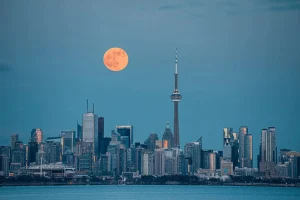 Toronto skyline at dusk with a full moon rising above the CN Tower, reflecting on the city's modern architecture.