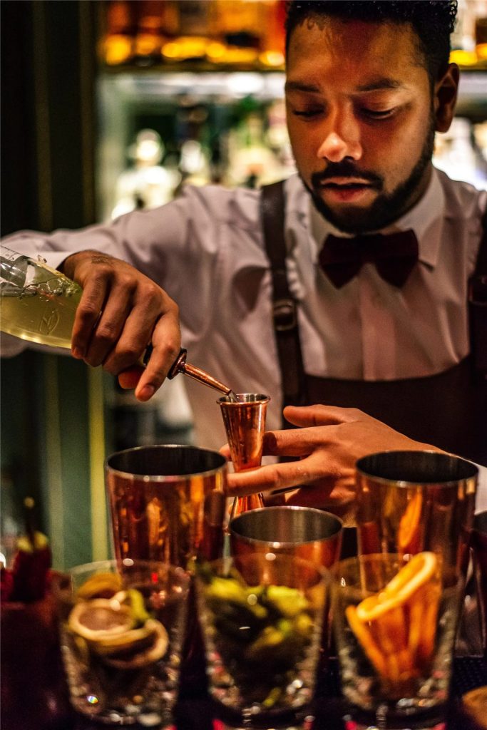 A bartender measuring a spirit into a copper jigger while preparing garnished cocktails at a bar