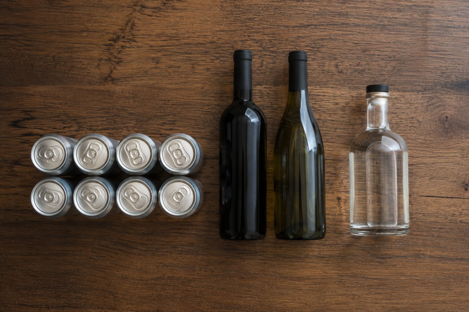 Eight beer cans, two wine bottles and one spirits bottle arranged on a wooden table to compare serving counts