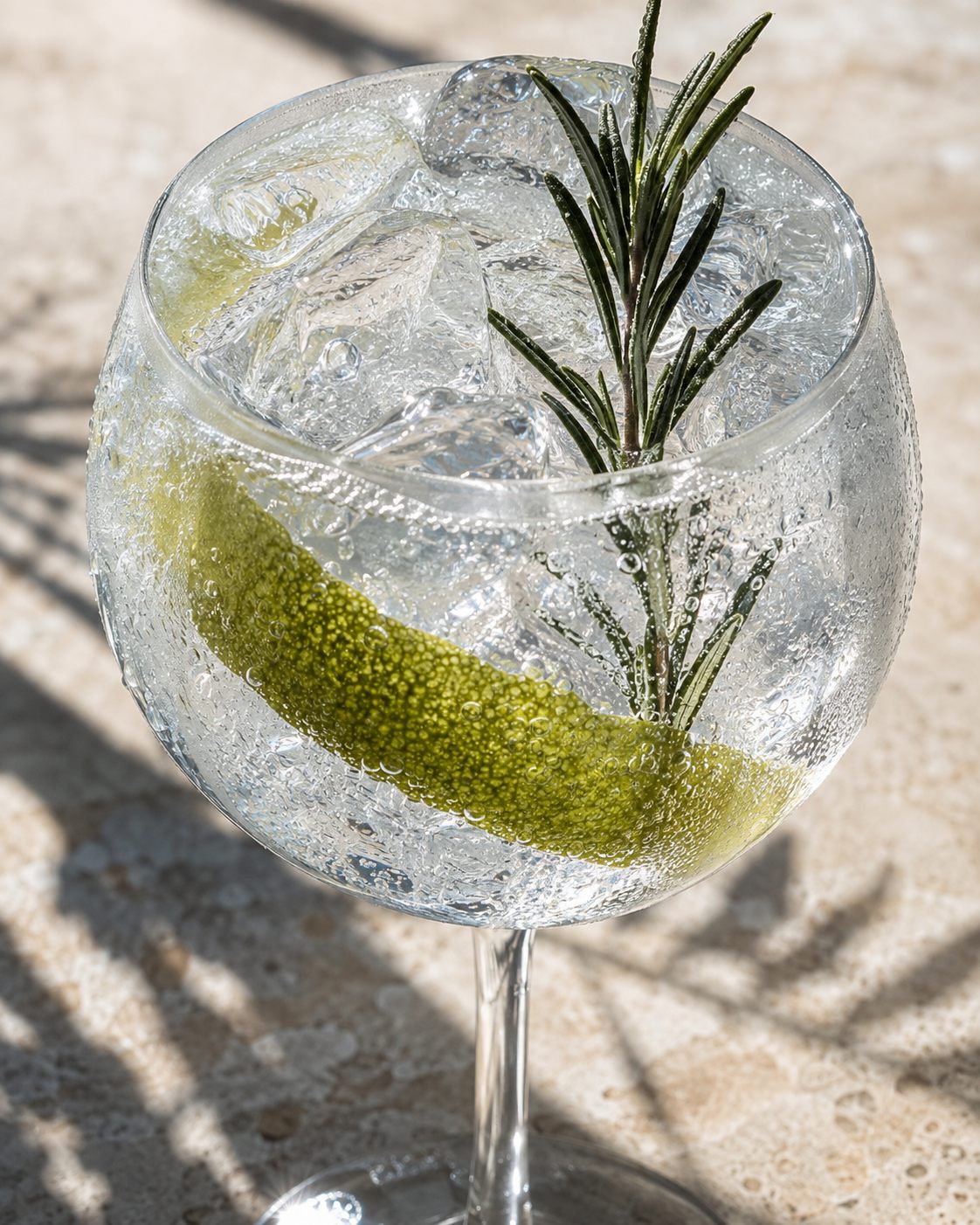 A gin and tonic in a balloon glass filled to the brim with ice, garnished with a wide lime peel and rosemary