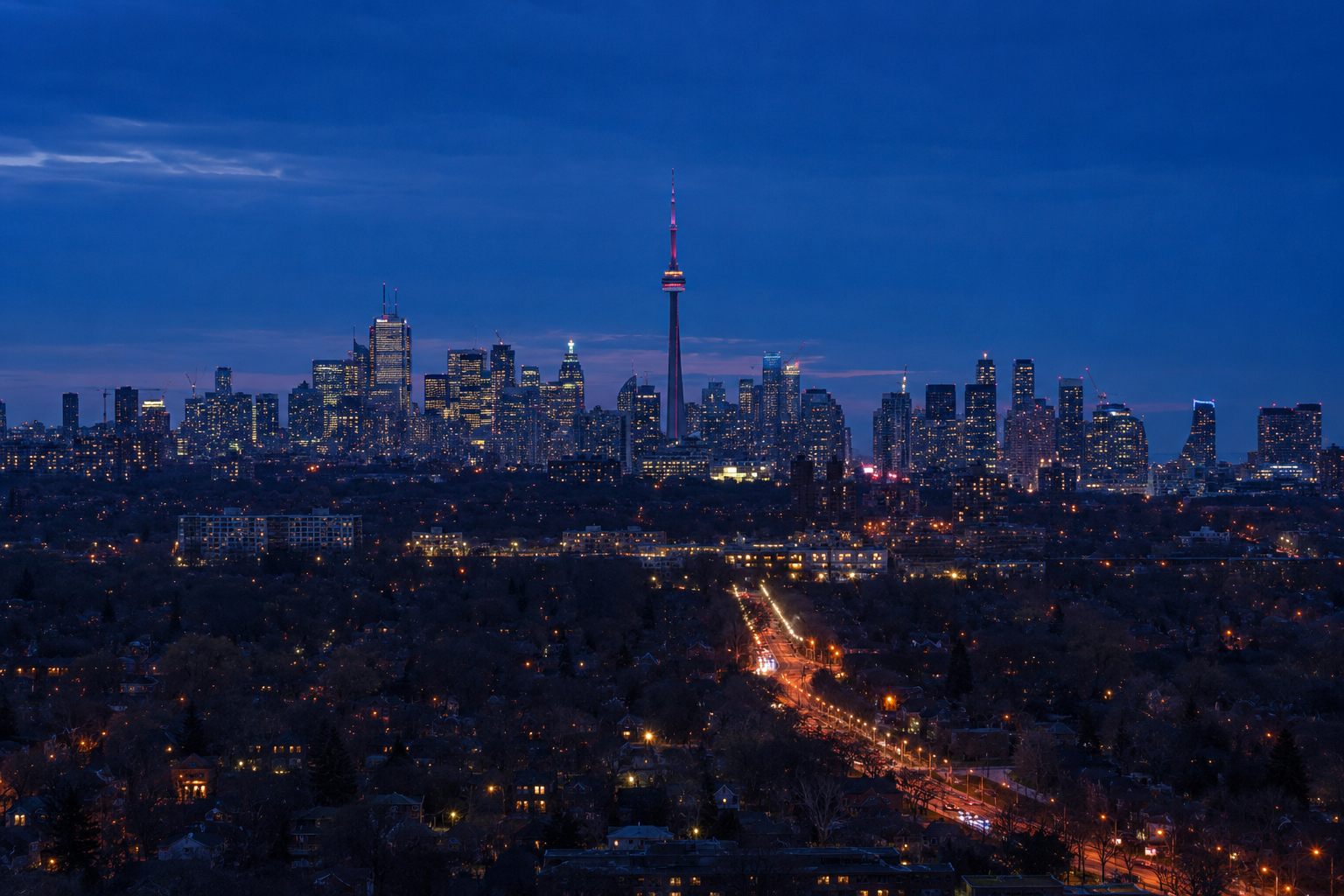 The Toronto skyline and CN Tower at blue hour, seen across low-rise residential suburbs with lit streets in the foreground